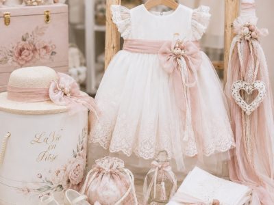 White dress for a young girl with blush pink sash and lace trim, displayed on a wooden rack with matching accessories in a pastel boutique scene.