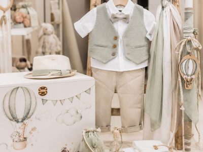 Toddler outfit on a wooden rack: white shirt, light green vest, beige shorts, and bow tie, surrounded by pastel decor and props.