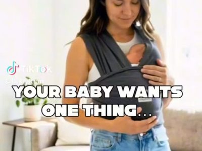 Mother wearing a baby wrap carrier, cradling a newborn against her chest in a bright living room, with the caption 'Your baby wants one thing...'.