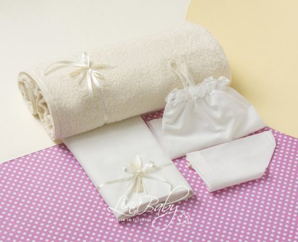 White baby linen set including a rolled towel, folded sheets, small drawstring bag, and a small fabric pouch arranged on a pink polka-dot surface.