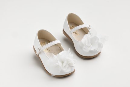 White Mary Jane-style children's shoes with a decorative white fabric flower and strap, photographed on a pale background for a formal look.