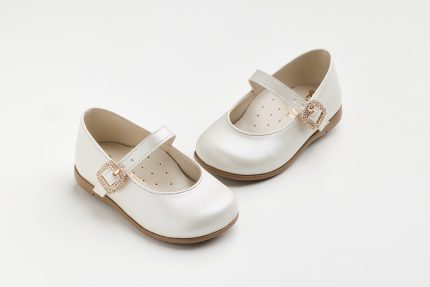 Pair of white children's Mary Jane shoes with decorative rhinestone buckles on a light brown sole against a white background.