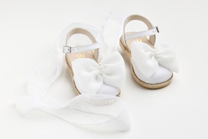 White baby sandals with ankle straps and large fabric bows, set on a white background with flowing ribbon