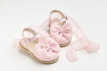 Pink children's sandals with large satin bows and ankle straps, placed on a white surface with pale pink fabric in the background.