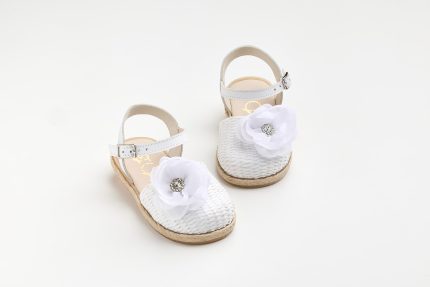 Pair of white woven sandals with ankle straps and large white fabric flowers adorned with rhinestones, displayed on a light background.