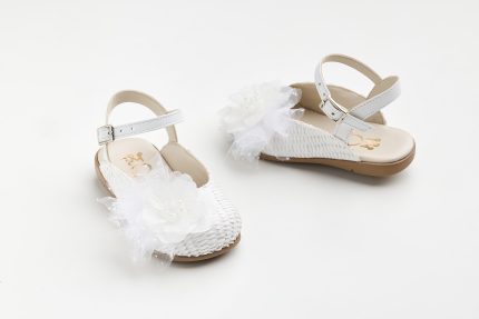White baby sandals with ankle straps and white fabric flower decorations on a light background behind them.