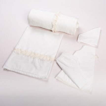 Set of white baby care items laid out on a pale pink background: towel, blanket, and matching clothing pieces with delicate trim.