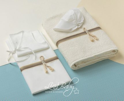 Baby bath set arranged on a striped blue mat: white onesie, bib, hat, and a folded towel with tan ribbons.