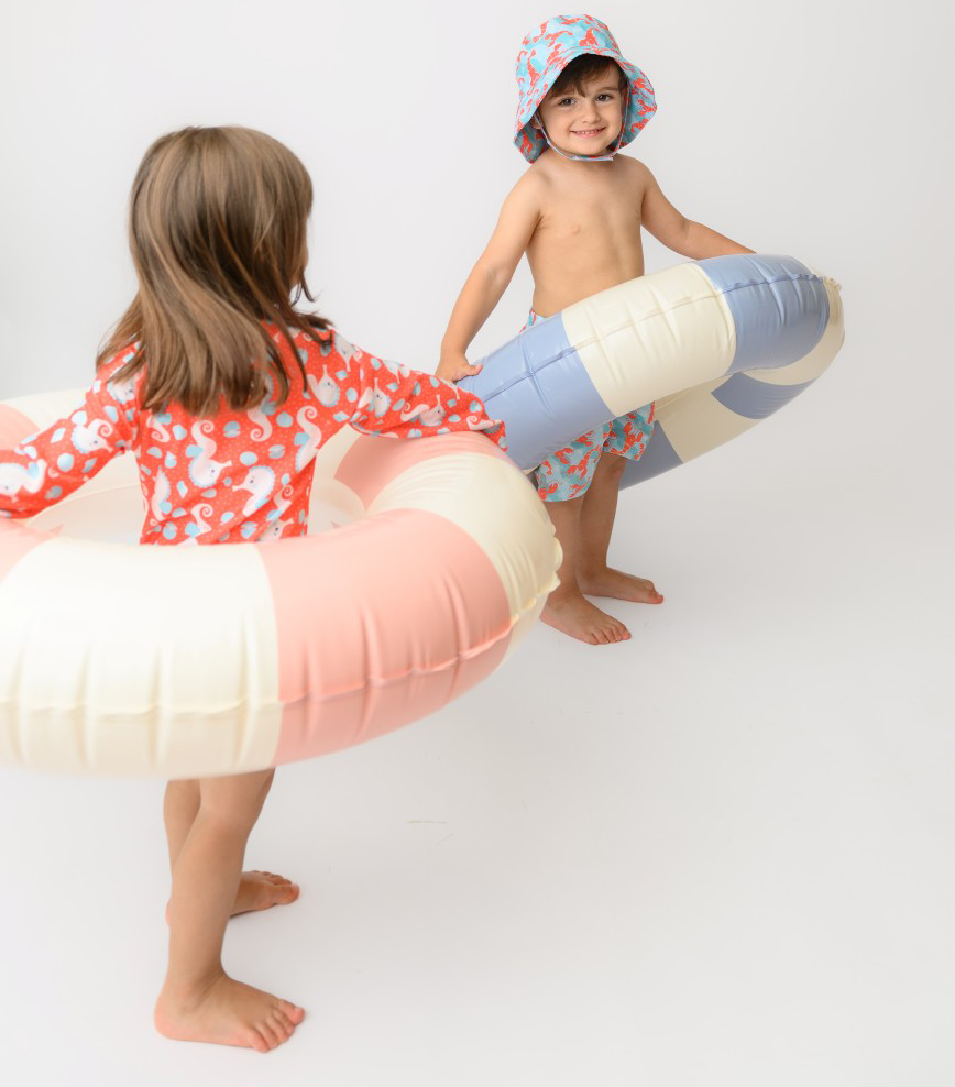 ZOO12604_12907_SS26_Swimsuit_SeahorseLobster_3 | La Vie en Rose Two young children in colorful swimsuits play with large inflatable inner tubes indoors on a white background, smiling and holding the rings.
