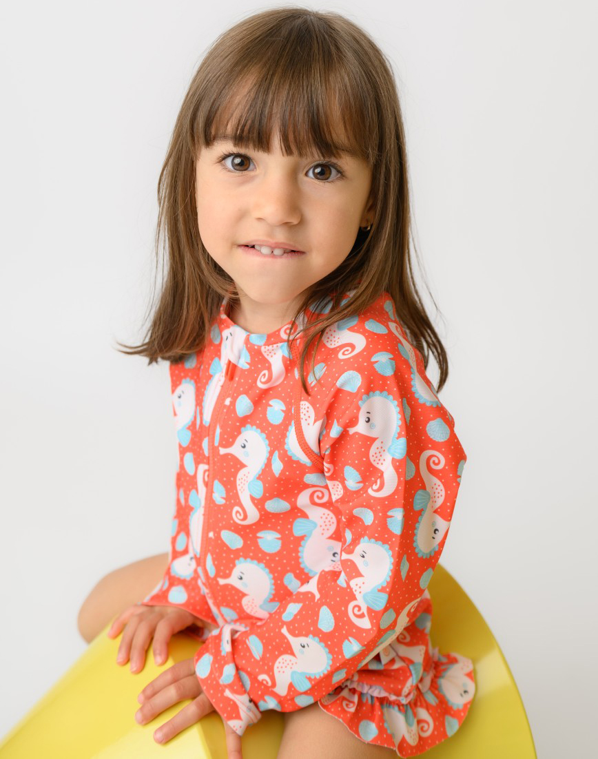 ZOO12604_SS26_OnepieceSwimsuit_Seahorse_2 | La Vie en Rose Young girl with brown hair in a red dress with sea horse pattern, sitting on a yellow chair against a white background.
