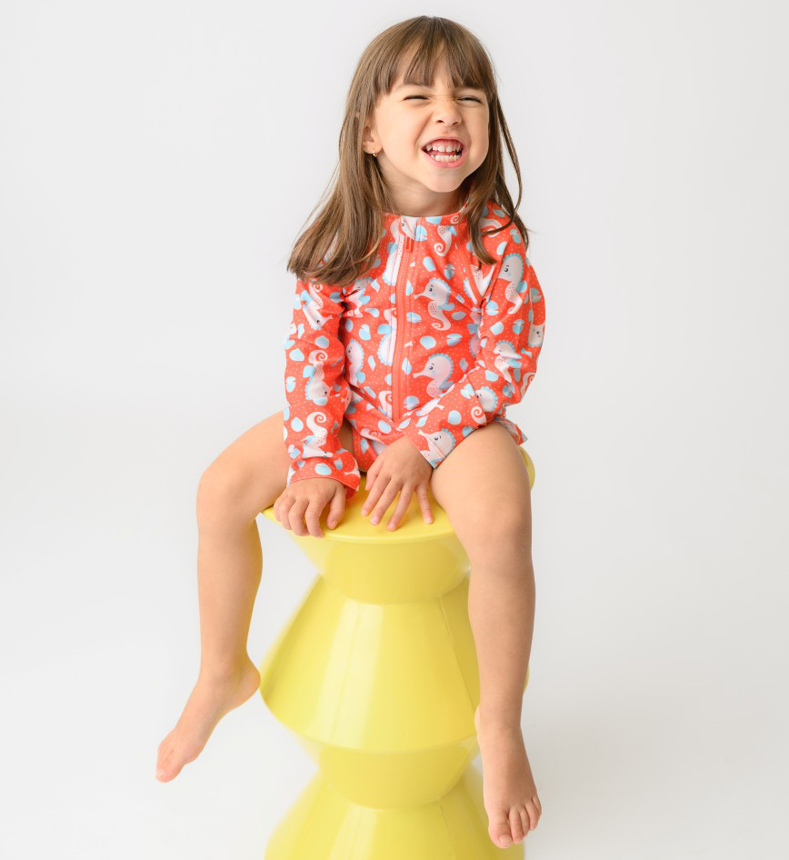 ZOO12604_SS26_OnepieceSwimsuit_Seahorse_3 | La Vie en Rose Smiling young girl in a red patterned swimsuit sits on a yellow stacked stool against a white studio backdrop.