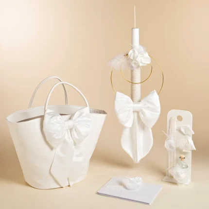 White wedding-themed décor: a white embossed tote bag with a large bow, a tall decorated candle with circular gold rings, a white bow and feather accents, and a small gift display with ribbons.