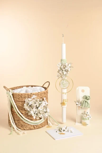 Beige background with a tall white candle adorned with flowers, a wicker basket of white blossoms, and a white decorative item with ribbons and floral accents.