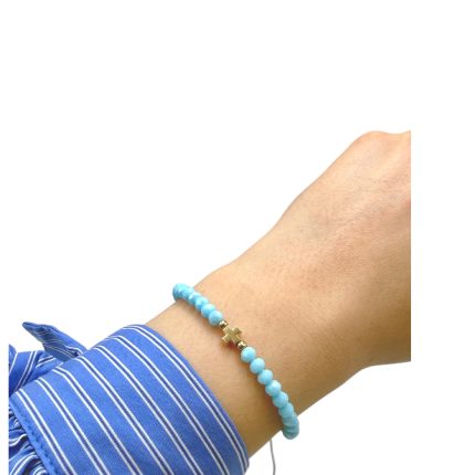 Light blue beaded bracelet with a small gold cross charm on a wrist with a blue striped cuff nearby.