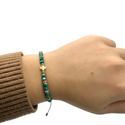 Teal beaded bracelet with a gold cross charm on a person's wrist leant against a brown knit sleeve.