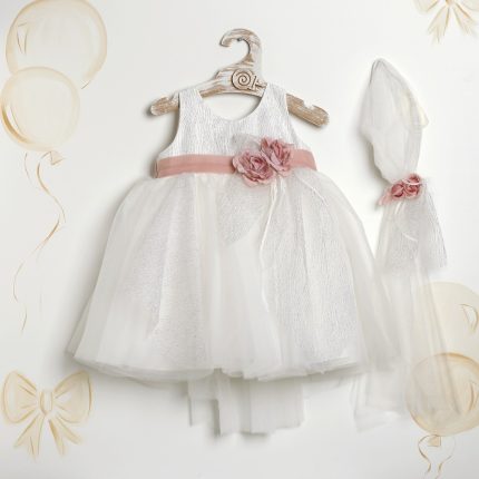 White baby dress with pink sash and fabric flower on a wooden hanger, plus a matching veil and headpiece, with pale balloon decor in the background.