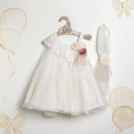 White baby dress with lace swirls and a ruffled neckline on a wooden hanger, accented by cream and pink flowers with a matching sheer veil beside it.