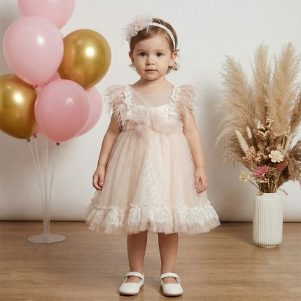 Toddler girl in a pale pink, lace-trim dress standing indoors with pink/gold balloons and a vase of dried flowers nearby.