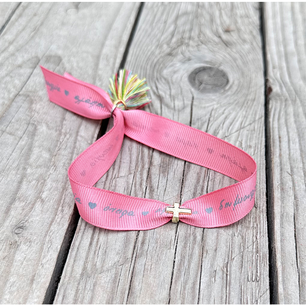 054040E0-833D-E745-C6F2-018C1B1D8FAB_11 | La Vie en Rose Pink fabric bracelet with a gold cross charm and a multicolor tassel on weathered wooden planks to the left of center.