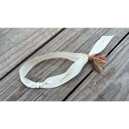 White fabric bracelet with a small gold cross charm and a multicolored tassel, placed on weathered wood.