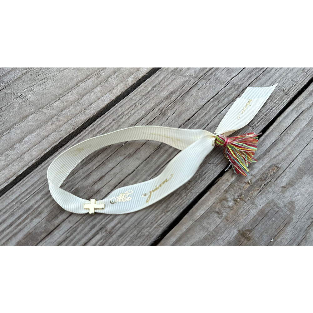0DFFC796-6AEC-6ABC-4291-018C1B758B09_11 | La Vie en Rose White fabric bracelet with a small gold cross charm and a multicolored tassel, placed on weathered wood.