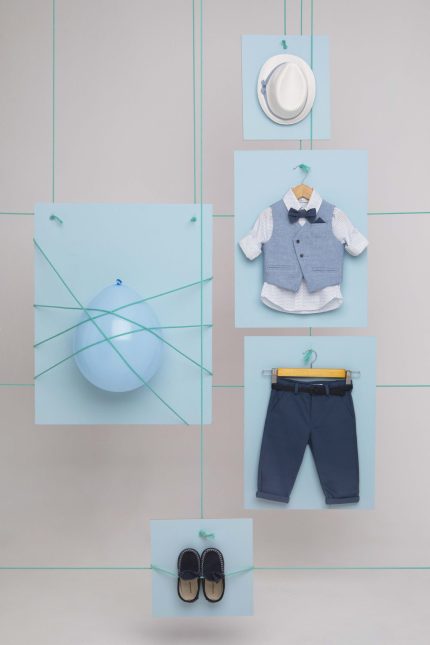 Flat lay of a toddler's formal outfit: light blue vest with bow tie, white shirt, navy pants, displayed on pale blue cards with strings.