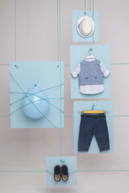Collage of pastel blue panels on a wall showing children’s clothing: a white hat, blue vest over a polka-dot shirt, navy pants, and matching shoes, all suspended by green cords.