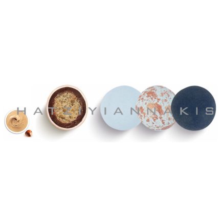Row of five round bath bombs in pastel to dark tones (light blue, speckled, and navy), with a small dish of creamy lotion on the far left.