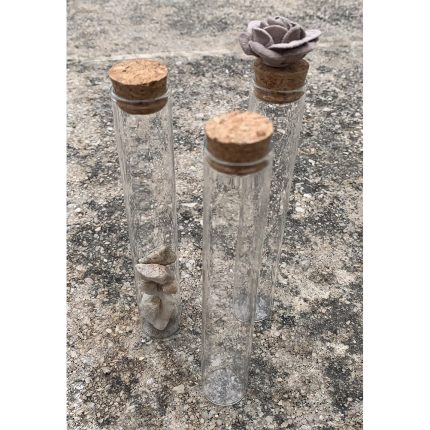 Three tall glass vials with cork stoppers stand on a rough outdoor surface; the left contains rocks, the rightmost has a fabric flower on top.