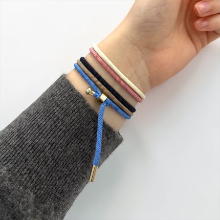 Woman's wrist with several colorful cord bracelets stacked, including blue adjustable cord with gold ends, worn over a gray knit sleeve.