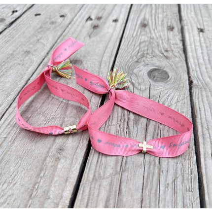 Pair of pink fabric bracelets with small gold cross charms and multicolored tassels on a weathered wooden surface.