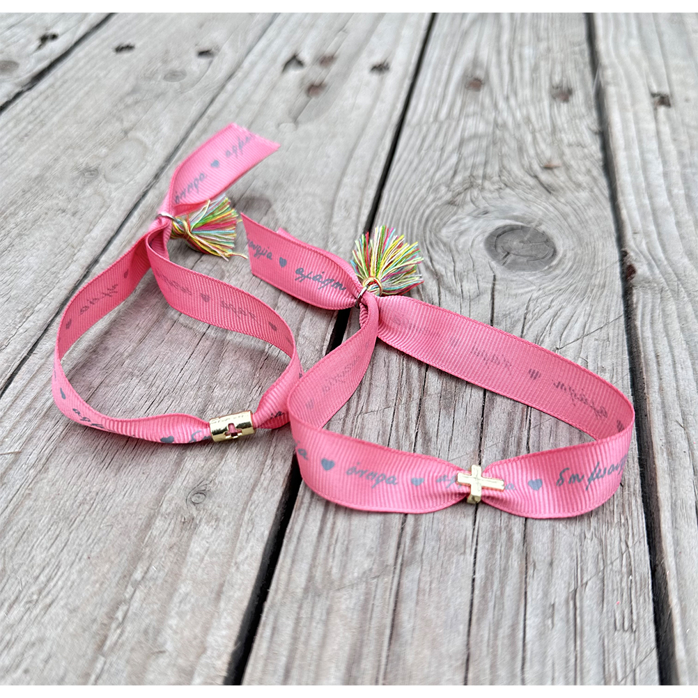 982EFD7E-25AB-ACA0-7DAA-018C1B1F20B0_21 | La Vie en Rose Pair of pink fabric bracelets with small gold cross charms and multicolored tassels on a weathered wooden surface.