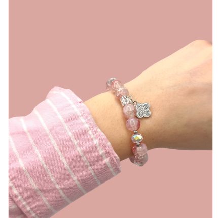Beaded pink bracelet with a silver clover charm on a model's wrist, pink striped shirt cuff visible against a pink background.
