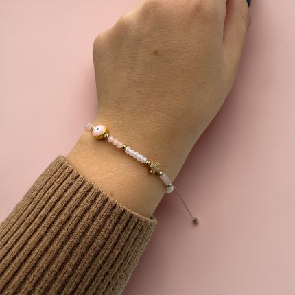 Wrist wearing a pink beaded bracelet with small gold accents and a smiling face bead against a pink background.