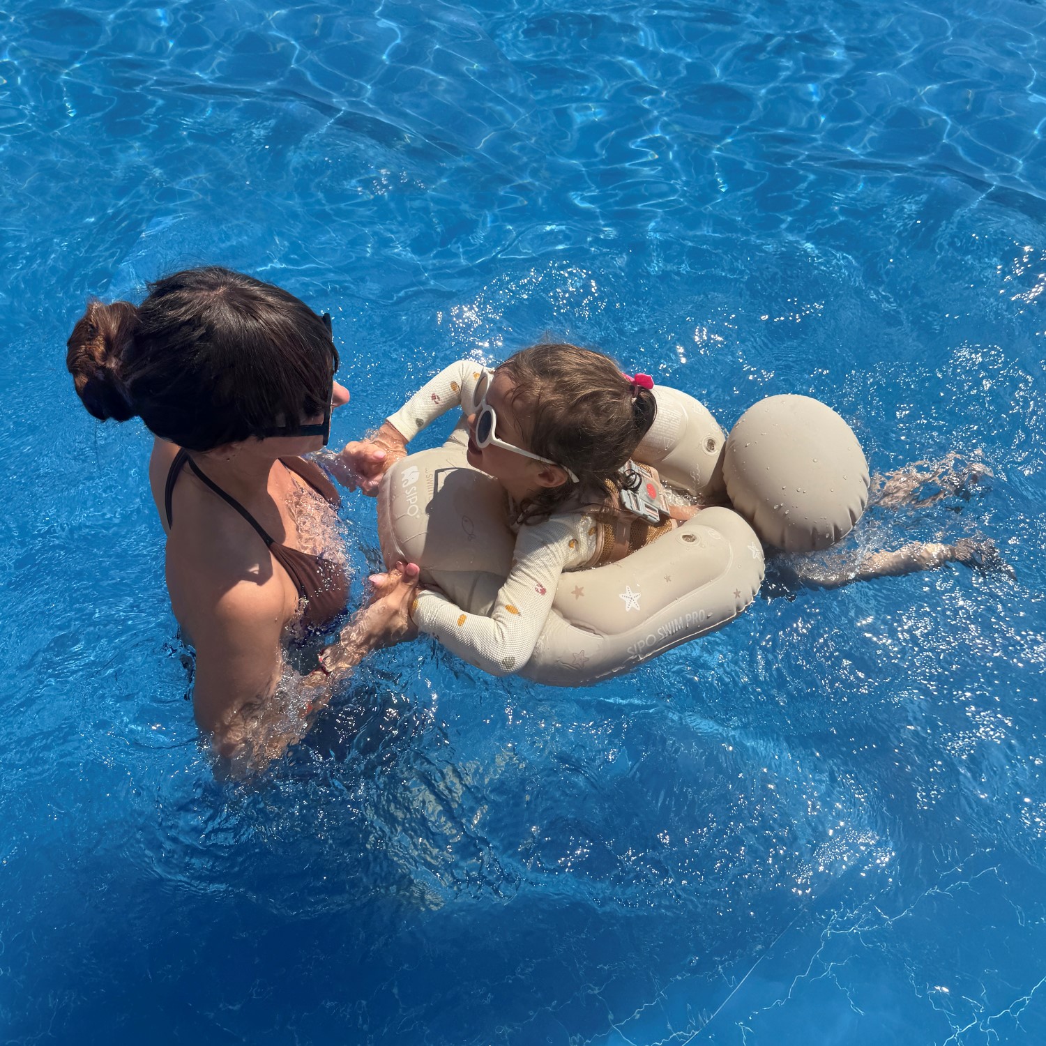 SIPO-SWIM-PRO_09 | La Vie en Rose Adult in a dark bikini helps a child seated in a beige inflatable swim float in a bright blue pool.