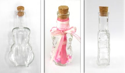 Three clear glass bottles with corks arranged in a row: a plain irregularly shaped bottle left, a center decorative bottle with pink ribbons and beads, and a tall rectangular bottle right.