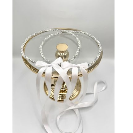 Gold-toned cake stand displaying two diamond-encrusted ring motifs with white satin ribbons. Used as a wedding ring display.