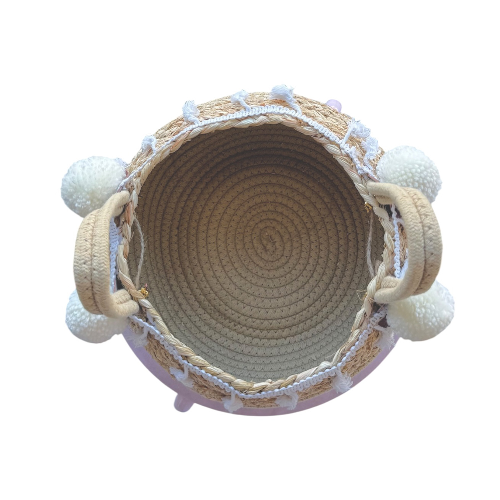 97035EE5-EB77-C919-6E1B-019DB0501FB5_21 | La Vie en Rose Top-down view of a round woven straw bag with braided rim, white pom-poms on the sides, and short handles.